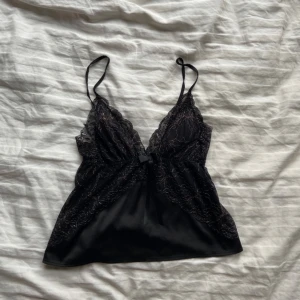Svart spetslinne från Censored - Elegant svart linne med spetsdetaljer och smala axelband från Censored. Linnet har en vacker spetsdesign över bysten och en liten rosett framtill. Perfekt för en stilfull look.
