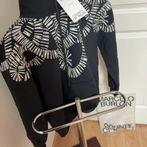 Svart hoodie från Marcelo Burlon  - Snygg svart hoodie från Marcelo Burlon med ett unikt vitt mönster över axlarna och bröstet. Tröjan har en klassisk huva och en praktisk magficka. Perfekt för en stilren och modern look.                            Skriv för info eller vid intresse!