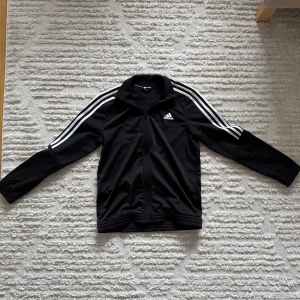 Adidas cardigan - Snygg svart cardigan från Adidas med de klassiska tre vita ränderna längs ärmarna. Jackan har en dragkedja framtill och Adidas-loggan på bröstet. Perfekt för sportiga aktiviteter eller en avslappnad stil.
