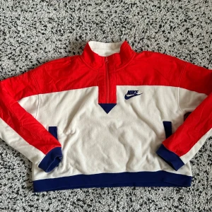 Nike fleece sweatshirt - Säljer en snygg tröja från Nike i rött och vitt med blå detaljer. Tröjan har en halv dragkedja och långa ärmar. Perfekt för en sportig look. Den är tillverkad i ett mjukt material som känns skönt mot huden.