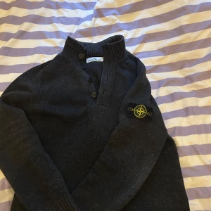 Svart tröja från Stone Island - Säljer en svart tröja från Stone Island med ribbad krage och knappar framtill. Tröjan har en ikonisk logotyp på ärmen och är perfekt för en stilren look. Passar bra till både jeans och chinos. Storlek M 