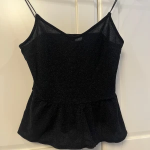 Svart glittrig topp - Snygg svart glittrig linne med smala axelband och en lätt utsvängd nederdel. Perfekt för en kväll ute eller en festlig tillställning. Enkel men elegant design som passar till många olika outfits.