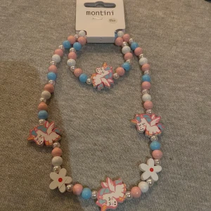 Färgglatt halsband och armband med enhörningar från Ur&penn - Charmigt set med halsband och armband från Montini. Smyckena har färgglada pärlor i rosa, blått och vitt, dekorerade med söta enhörningar och blommor. Perfekt för att lägga till en lekfull touch till din outfit!