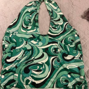 Grön mönstrad halterneck topp - Snygg grön halterneck topp med ett unikt virvlande mönster i olika gröna nyanser och svart. Perfekt för en somrig look med sin ärmlösa design och djupa urringning.