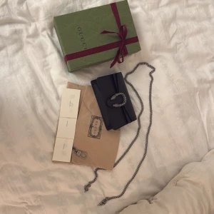 Svart väska från Gucci - Säljer en elegant svart axelväska från Gucci med en dekorativ metallspänne framtill. Väskan har en kedjerem och är tillverkad i skinn. Perfekt för att ge en lyxig touch till din snygga outfit. Ny pris är 11,750kr. Då den är helt oanvänd så vill jag helst ha 10 000 för den. 