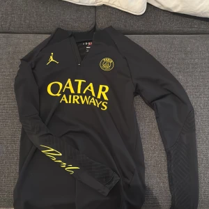 Svart långärmad fotbollströja från Jordan x PSG - Säljer en svart långärmad fotbollströja från Jordan i samarbete med Paris Saint-Germain. Tröjan har gula detaljer inklusive Qatar Airways-loggan på bröstet och en gul Jordan-logga. Den har en halv dragkedja vid halsen och är i slim fit-modell. Perfekt för fotbollsfans som vill ha något unikt och stilrent!