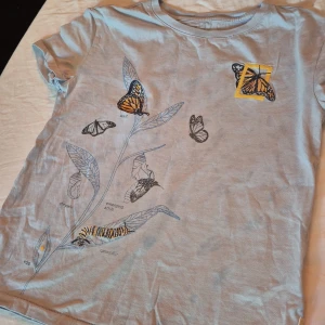 Grå t-shirt med fjärilsmotiv från GAP Kids x National Geographic - Säljer en superfin grå t-shirt från GAP Kids i samarbete med National Geographic. Den har ett coolt fjärilsmotiv på framsidan och texten 'SPREAD YOUR WINGS' i gult på baksidan. Gjord av 100% ekologisk bomull, så den är både bekväm och miljövänlig. Perfekt för sommaren! 🦋