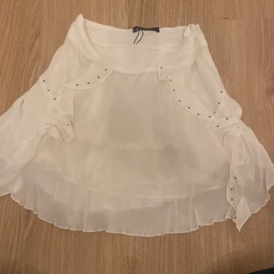 Vit kjol från Zara - Säljer en superfin vit kjol från Zara i storlek S. Kjolen är gjord av ett lätt och luftigt material, perfekt för sommaren. Den har en volangdetalj och små silverfärgade pärlor som ger den en extra touch. Passar perfekt till både vardag och fest!