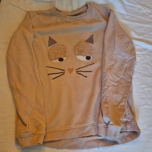 Beige tröja med kattmotiv - Säljer en supergullig beige tröja med ett kattansikte på framsidan. Tröjan har långa ärmar och är perfekt för höst och vinter. Den är i bra skick och väldigt mysig att ha på sig. Perfekt för kattälskare! 🐱