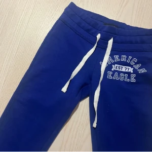 Blå mjukisbyxor från American Eagle - Säljer ett par superbekväma blå mjukisbyxor från American Eagle. De har en vit snörning i midjan och texten 'American Eagle EST 77' tryckt på vänster höft. Perfekta för en chill dag hemma eller en snabb tur till affären. De är i storlek S och sitter lågt på höften. Passar perfekt till en avslappnad stil!