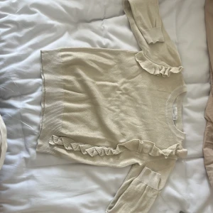 Beige stickad tröja med volanger - Säljer en superfin beige/guldig stickad tröja med volanger på ärmarna och längs sidorna. Tröjan är långärmad och har ribbstickade muddar vid ärmslut och nederkant. Perfekt för höst och vinter! 🧡