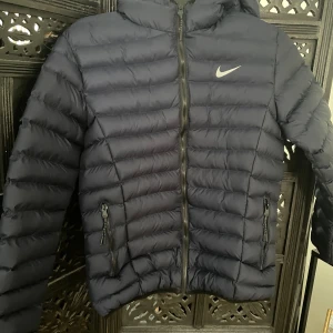 Nike puffer jacket - Marinblå puffer jacka fr Nike.  Den är i stl L för barn, så passar 14-16 år.  Den är i jättefint skick, dock är det ett hål på undersidan av luvan. Detta kan lätt sys 
