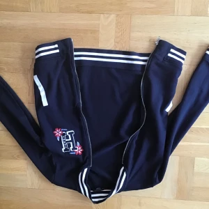 Svart bomberjacka från Tommy Hilfiger - Säljer en snygg svart kofta från Tommy Hilfiger. Jackan har vita detaljer som ränder på kragen, ärmsluten och nederkanten. Den har också en dragkedja framtill och ett broderat H med blommor på ena sidan. Perfekt för vår och höst! Skriv gärna om ni bar några frågor 💕