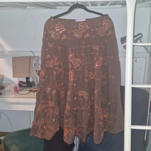 Brun kjol med broderade blommor - Charmig brun kjol med vackra orange broderade blommor. Kjolen har en volangdesign som ger den en lekfull känsla. Perfekt för våren, tyvärr för kort för mig då den går till mitten av mina smalben (jag är 175cm).