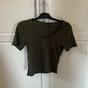 Olivgrön ribbad topp från H&M - Säljer en olivgrön ribbad topp från Veromoda med korta ärmar och knappar framtill. Perfekt för en avslappnad stil. Toppen har en rund halsringning och är i ett mjukt material.