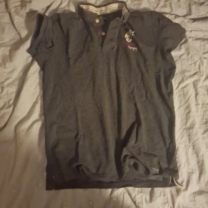 Mörkgrå pikétröja från Peak Performance - Säljer en stilren mörkgrå pikétröja från Peak Performance med broderad logga på bröstet. Tröjan har korta ärmar och en klassisk krage med knappar. Perfekt för en avslappnad look.