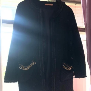 Svart kappa med kedjedetaljer - Elegant svart kappa med långa ärmar och stiliga kedjedetaljer vid fickorna. Perfekt för en chic look. Den har en klassisk design som passar till många tillfällen.