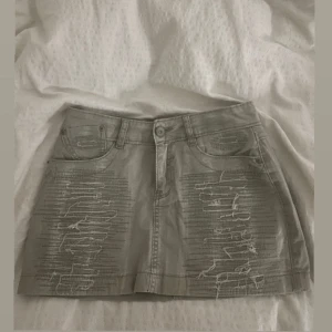 Grå kortkjol med slitningar - Snygg grå kortkjol med slitna detaljer och femficksdesign. Kjolen har en knapp och dragkedja framtill samt en cool patch på baksidan. Perfekt för en avslappnad stil.