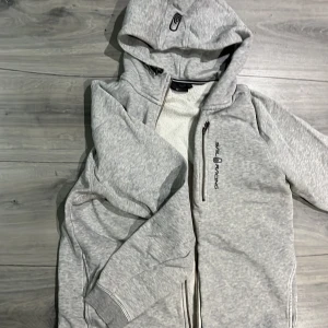 Grå huvtröja från Sail Racing - Säljer en grå huvtröja från Sail Racing med dragkedja och broderad logga på bröstet samt luvan. Tröjan har långa ärmar och en praktisk huva. Har lite färg missbildning på vänster armbåge men inget som syns så mycket. 