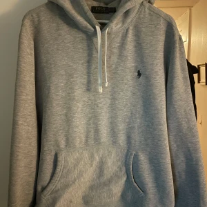 Grå hoodie från Polo Ralph Lauren - Säljer en stilren grå hoodie från Polo Ralph Lauren med klassisk känguruficka och justerbar huva. Den har en liten broderad logga på bröstet och är perfekt för en avslappnad look. Passar bra till både jeans och joggers. Har en liten mörk fläck som ni ser på bild 4, dock inget jag någonsin stört mig på.
