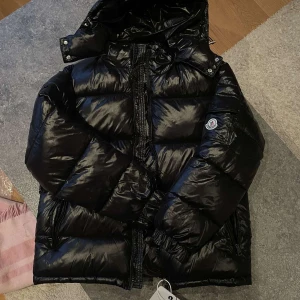 Svart dunjacka från Moncler - Säljer en snygg svart dunjacka från Moncler. Jackan har en glansig finish och är utrustad med dragkedja och huva. Perfekt för kalla dagar med sin varma och puffiga design. Moncler-logga på ärmen ger en exklusiv touch.