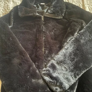 Svart teddyjacka från Lindex - Mysig svart teddyjacka från Lindex i storlek 164. Jackan har en dragkedja framtill och är perfekt för kyligare dagar. Den är tillverkad i ett mjukt och fluffigt material som ger en skön känsla.