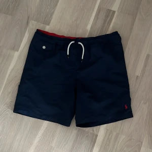 Ralph Lauren badbyxor  - Snygga marinblå badshorts från Ralph Lauren med röd insida och vit snörning i midjan. Shortsen har en liten broderad logga i rött på benet och en knappdetalj på fickan. Perfekta för sommarens stranddagar!