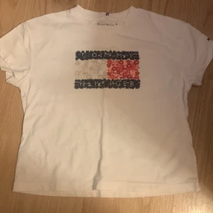 Vit t-shirt från Tommy Hilfiger - Säljer en vit t-shirt från Tommy Hilfiger med ett ikoniskt märke i rött, vitt och blått på framsidan. T-shirten är kortärmad och har en klassisk passform. Perfekt för en stilren och avslappnad look.