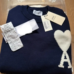 Ami paris - Säljer en snygg mörkblå tröja från Ami med ett vitt hjärta och bokstaven A på bröstet. Tröjan har rund hals och långa ärmar, perfekt för en stilren look. Den är tillverkad i ett mjukt material som känns skönt mot huden.