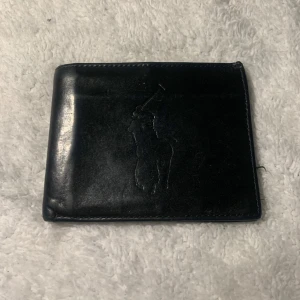 Svart plånbok från Ralph Lauren - Snygg svart plånbok i skinn från Ralph Lauren med präglad logotyp på framsidan. Plånboken har flera kortfack och en sedelficka, perfekt för att hålla ordning på dina kort och kontanter.