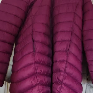 Vinröd dunjacka - Säljer en snygg vinröd dunjacka med quiltad design. Jackan har långa ärmar och en dragkedja framtill. Perfekt för kyliga dagar och ger en stilren look. Passar bra till både vardag och utflykter.