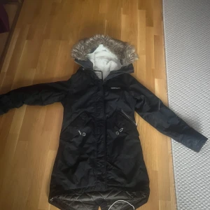 Svart parkajacka från Didriksons - Säljer en svart parkajacka från Didriksons med mysigt foder och pälskantad huva. Jackan har dragkedja och knappar framtill samt praktiska fickor. Perfekt för kyliga dagar!