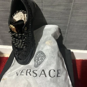 Svarta sneakers från Versace med gulddetaljer - Snygga svarta sneakers från Versace med iögonfallande gulddetaljer. Skorna har snörning och ett mönster med Versace-logotypen. Perfekta för den som vill ha en lyxig touch i sin skogarderob.