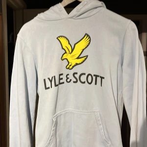 Ljusblå hoodie från Lyle & Scott - Säljer en ljusblå hoodie från Lyle & Scott med deras ikoniska gula örn och logga på framsidan. Tröjan har en klassisk känguruficka och en bekväm huva. Perfekt för en avslappnad stil. Strl 15-16 år