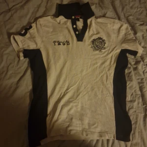 Vit och svart pikétröja från U.S. Polo Assn - Storlek M men kan även passa L. Priset kan variera. Säljer en stilren vit pikétröja från Ralph Lauren med svarta detaljer och broderad logga. Tröjan har korta ärmar och en klassisk krage med knappar. Perfekt för en avslappnad men snygg look.