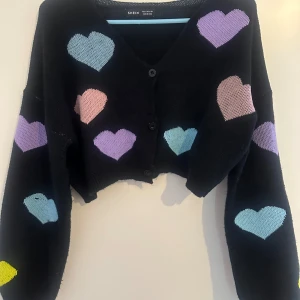 Svart kofta med hjärtmönster från Shein - Säljer en svart kofta från Shein med söta hjärtan i rosa och lila. Perfekt för att ge en färgglad touch till din outfit. Koftan har långa ärmar och en bekväm passform.