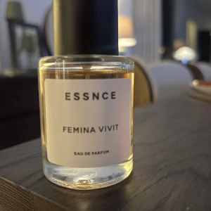 Essnce Femina Vivit Eau de Parfum - Elegant parfymflaska från Essnce med doften Femina Vivit. Flaskan är genomskinlig med en enkel vit etikett och svart kork. Perfekt för den som söker en sofistikerad och tidlös doft.