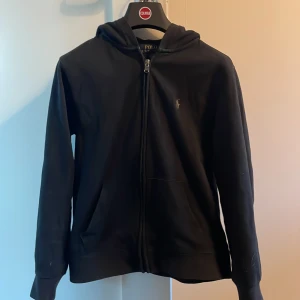 Svart hoodie från Ralph Lauren - Säljer en stilren svart hoodie från Ralph Lauren med dragkedja och broderad logga på bröstet. Perfekt för en avslappnad look. Den har långa ärmar och en praktisk huva. Storlek L (barn) (dock liten i storlek, passar någon som är ca 150cm) Pris går att diskutera 