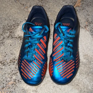 Adidas - Snygga fotbollsskor från Adidas i en cool blå och röd färgkombination. Skorna har snörning och vita detaljer på hälen. Perfekta för fotbollsplanen med sin stilrena design.