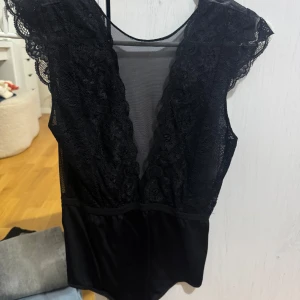 Svart spetsbody - Snygg svart body med spetsdetaljer och djup v-ringning. Den är ärmlös och har en elegant design som passar perfekt för en kväll ute. Materialet är mjukt och stretchigt för en bekväm passform.