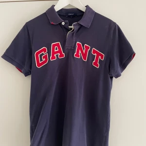 Mörkblå pikétröja från GANT - Säljer en snygg mörkblå pikétröja från GANT med röd och vit logga på bröstet. 