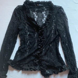 vintage spetsskjorta - Svart skjorta i spets med volanger vid urringningen, ärmarna och nedtill. Med knappar. Jättefin och trendig💋Passar M-L  Mått: Längd: 53cm Bredd axlar: 42cm Bredd armhåla till armhåla: 40cm