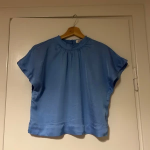 Blå blus från Rodebjer - Snygg blå blus från Rodebjer i ett mjukt och glansigt material. Blusen har en rund hals och korta ärmar. Inköpspris ca 2500-3000 kr. 