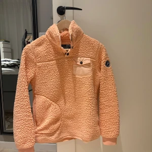 Orange fleecetröja från Pelle P - Mysig orange fleecetröja från Pelle P med hög krage och en knappförsedd bröstficka. Tröjan har långa ärmar och en broderad logga på ärmen. Perfekt för kyliga dagar. Tröjan är lite sliten långt med nedersta kanten (se bild), annars i gott skick!