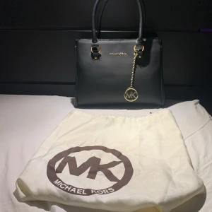 Svart handväska från Michael Kors - Snygg svart handväska från Michael Kors i skinn med gulddetaljer. Väskan har ett elegant hängsmycke med MK-logga. Dragkedjan är trasig men övriga väskan är i fint skick, men skulle alltså behöva lagas, därav priset. Hör av dig vid funderingar💕