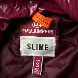 Vinröd dunjacka från Parajumpers - Säljer en snygg vinröd dunjacka från Parajumpers. Jackan har en quiltad design med dragkedja och en hög krage. Perfekt för kyliga dagar och ger en stilren look. Märkeslogga på ärmen ger en extra touch.