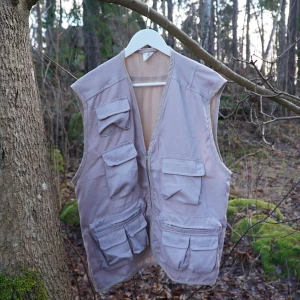 Beige väst med många fickor - Säljer en beige väst med flera praktiska fickor och dragkedja framtill. Perfekt för utomhusaktiviteter och har en avslappnad passform. Västen är tillverkad i ett slitstarkt material och har en neutral färg som passar till det mesta.