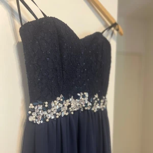 Mörkblå klänning med spets och pärldetaljer - Elegant mörkblå klänning med spetsöverdel och vackra pärldetaljer i midjan. Axelbandslös design med smala axelband och en flytande kjol. Perfekt för speciella tillfällen.
