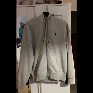 Grå hoodie från Ralph Lauren - Säljer en stilren grå hoodie från Ralph Lauren med dragkedja och huva. Tröjan har långa ärmar och en liten logga på bröstet. Perfekt för en avslappnad look.