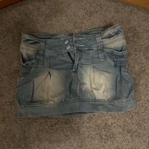 Blå jeanskjol - Snygg blå jeanskjol med två knappar och dragkedja framtill. Kjolen har smockdetaljer på sidorna och en kort längd som ger en trendig look. Perfekt för en avslappnad stil.